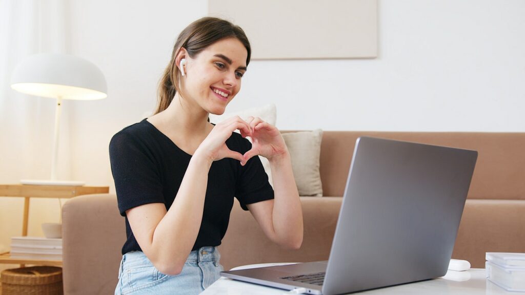 young woman showing heart symbol to team on virtual call with laptop