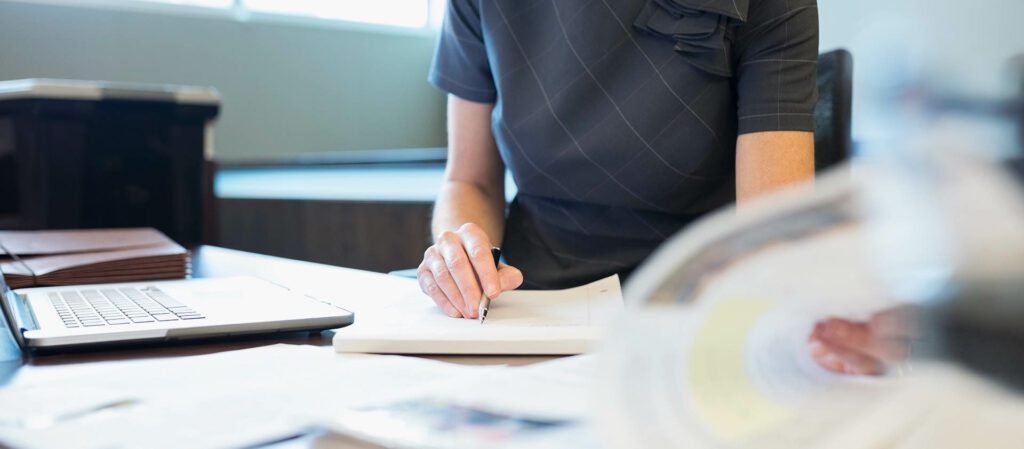 female employee writing notes while working with laptop from office desk.