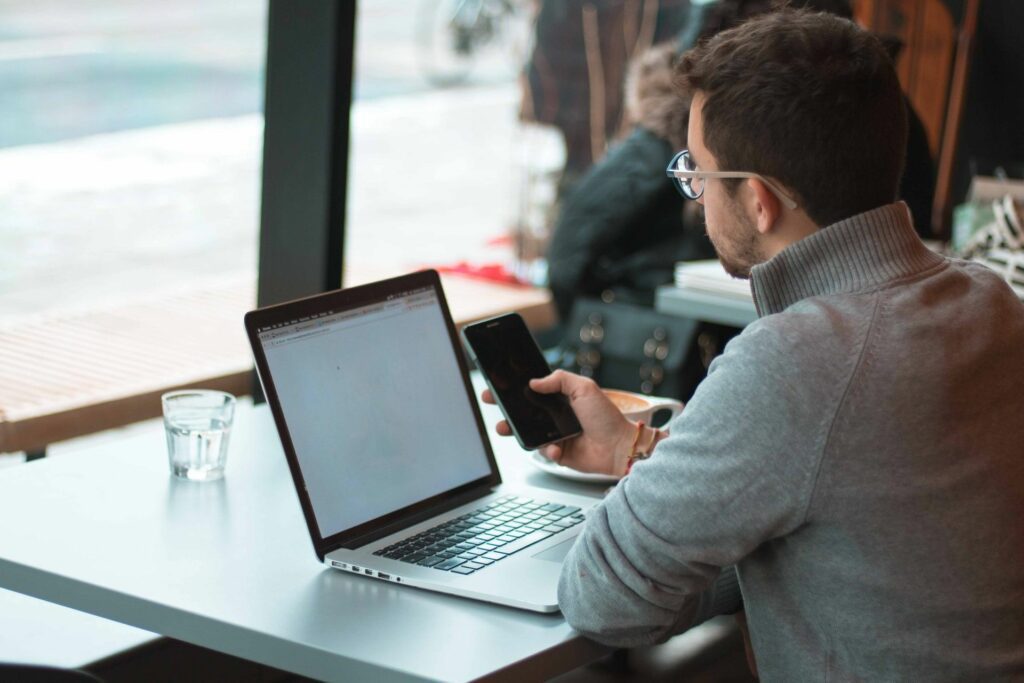 male employee using mobile while working with laptop from cafe