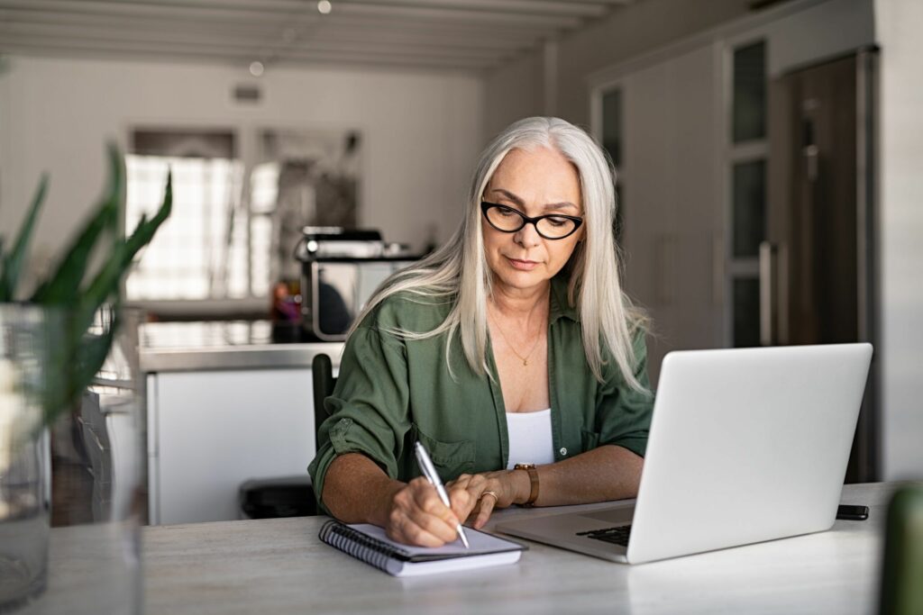 a woman writing on the notepad while working with laptop from home