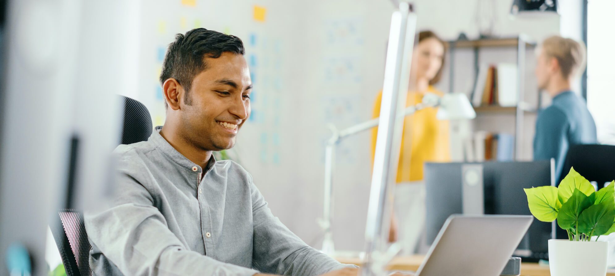 Handsome Smiling Indian Office Worker Sitting at His Desk works on a Laptop. In the Background Modern Office with Diverse Team of Young Professionals Working.