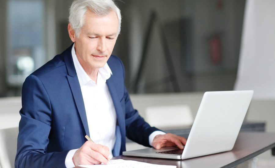 Senior Businessman dressed in blue suit writing notes while working on laptop.